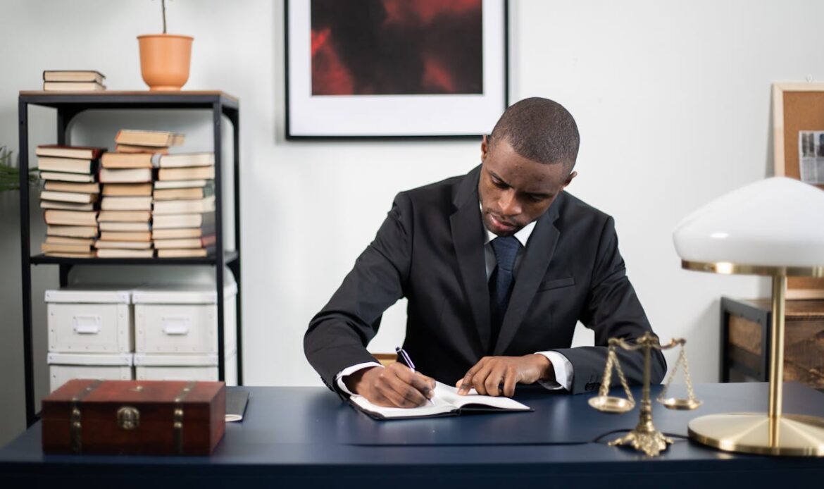 Businessman in formal suit writing notes at office desk with books and scales of justice.